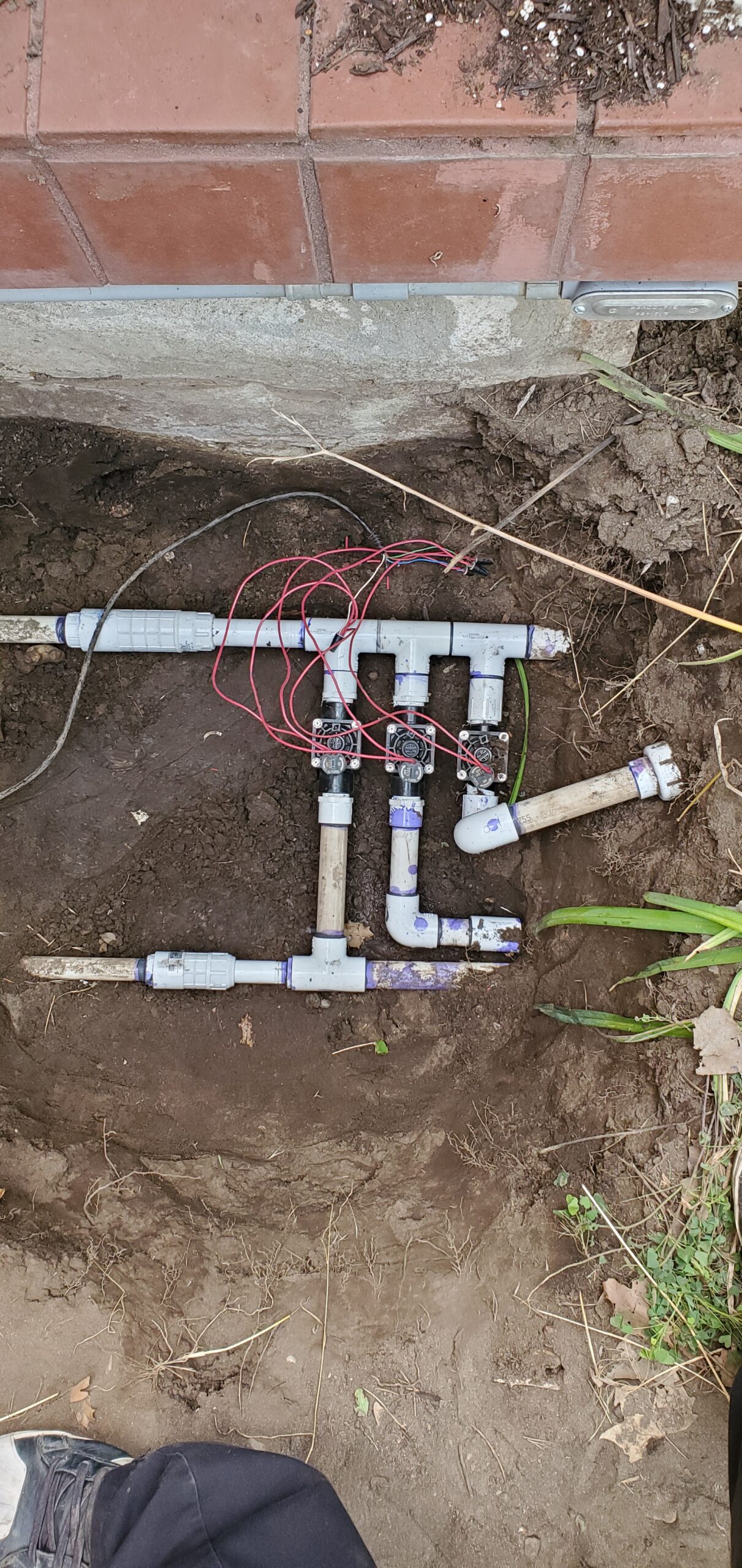 Exposed PVC irrigation pipe assembly with valves and wiring in a trench beside a brick wall