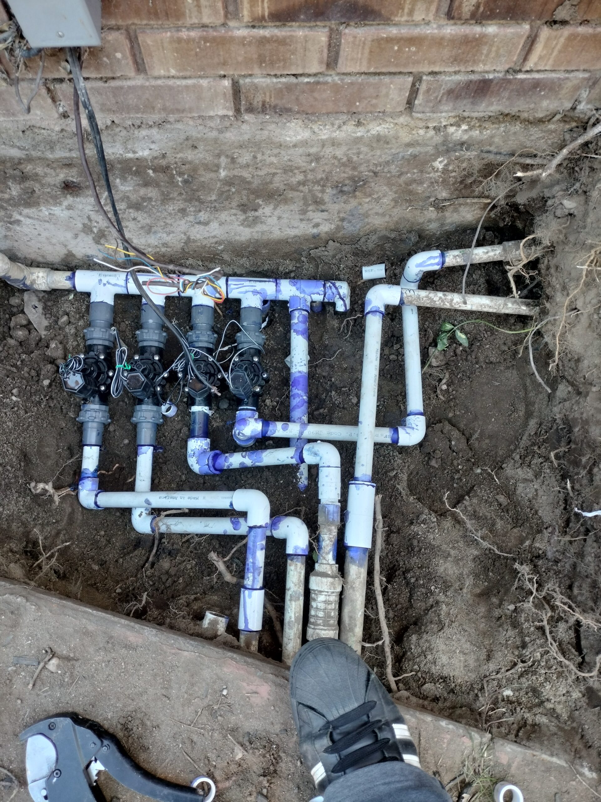 Exposed PVC irrigation pipe assembly with valves and wiring in a trench beside a brick wall